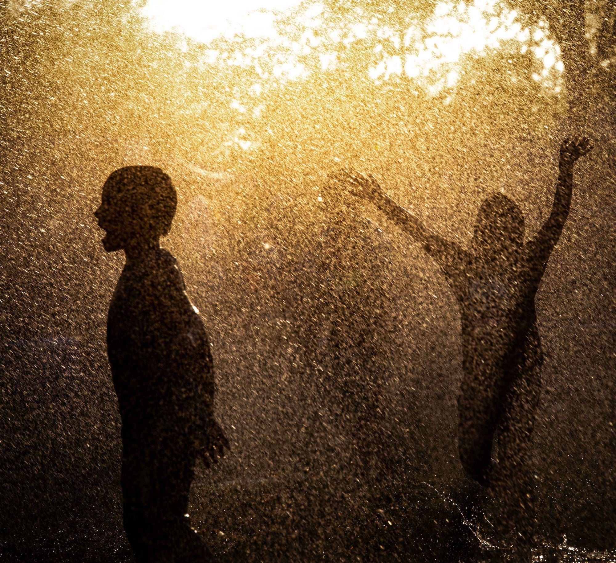 Children play outside in a splash pad, illuminated by the sun's rays shining through the water droplets.