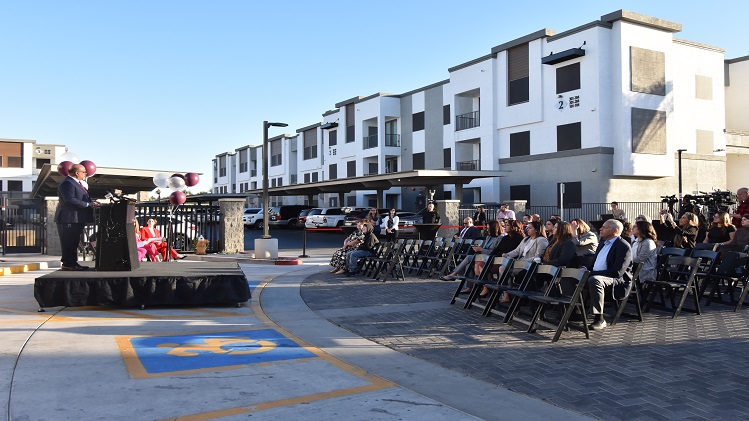 A crowd of people sit in chairs outside as a speaker addresses them from a podium at a press event.