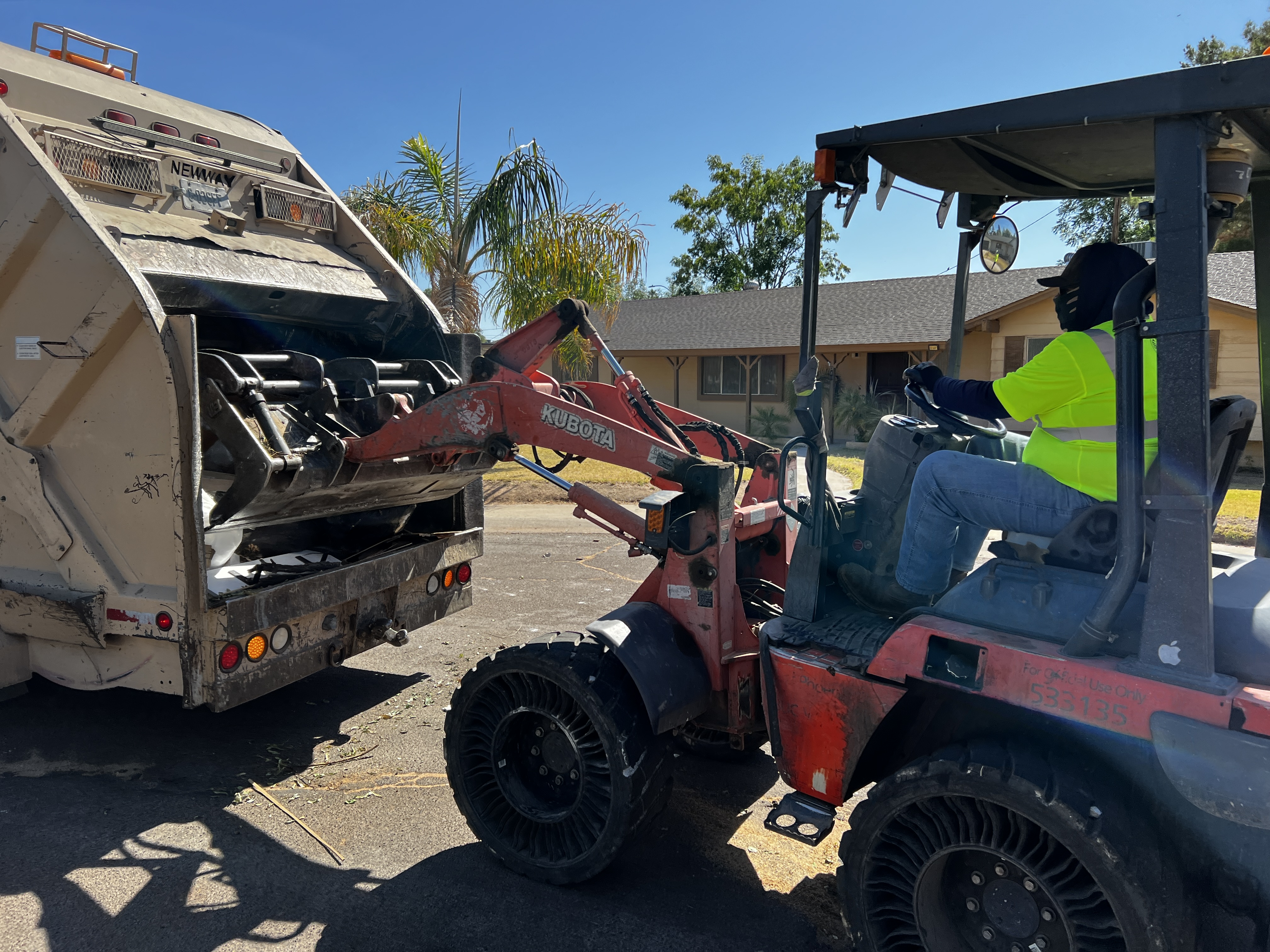 A bulk trash equipment operator uses a tractor to lift large items into a rear-loader garbage truck.